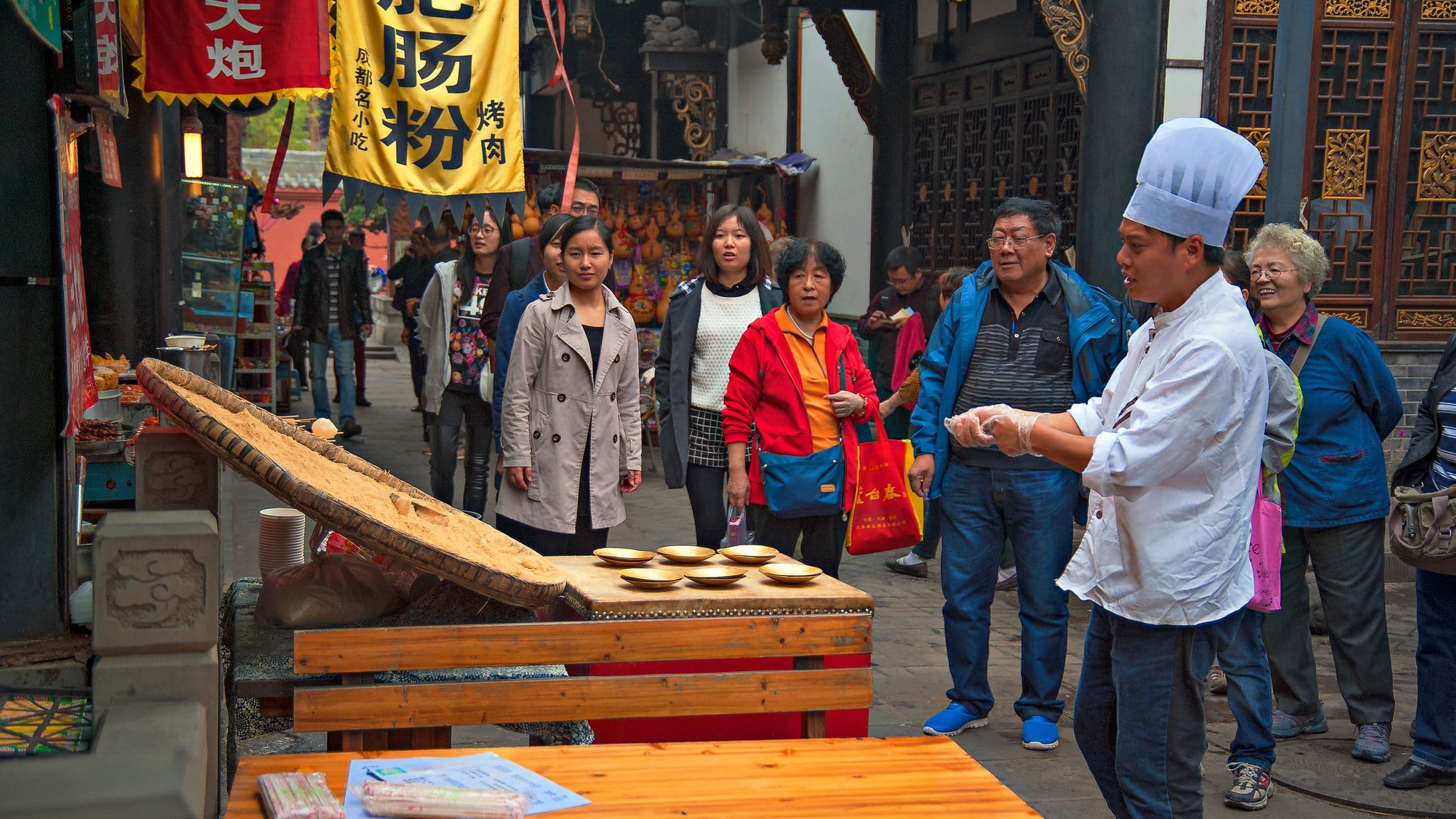 A chef preparing food in an open market, surrounded by visitors